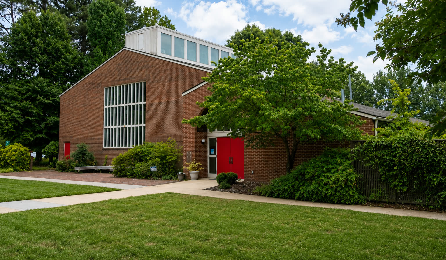 The courtyard entrance in summer: red doors under a brick gable, a shade tree beside them, and mown lawn crossed by a path